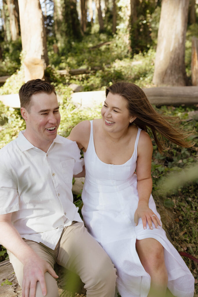 Laughing couple sitting on a fallen log during their San Francisco engagement session in the Presidio forest