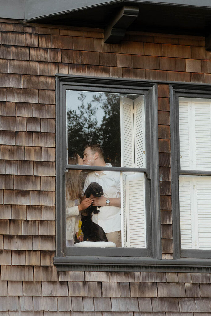 Engaged couple holding their cats inside an apartment in San Fransisco kissing through a window