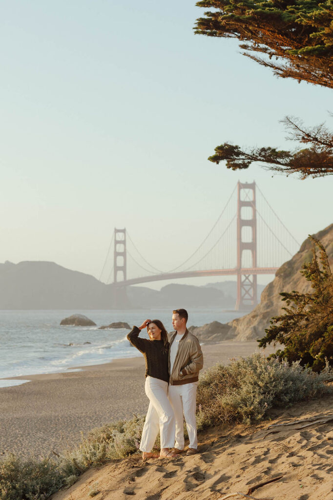 Engaged couple looking at the sunset barefoot on the beach with the Golden Gate bridge in the background