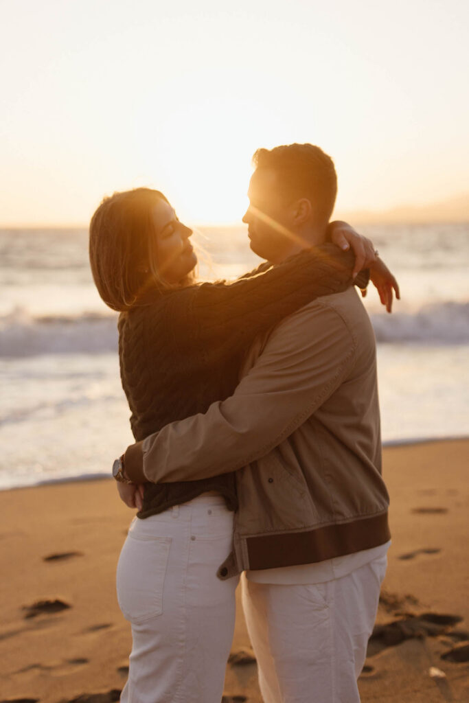 Engaged couple hugging while the sunset creates a sun flare on the beach in San Fransisco
