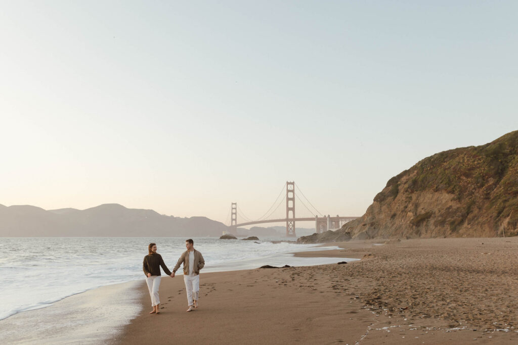 Landscape view of an engaged couple walking on the beach in San Fransisco at sunset with the Golden Gate bridge in the background