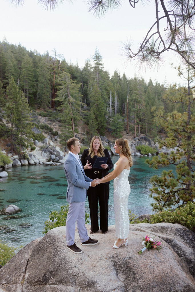 Elopement couple holding hands during ceremony in Lake Tahoe while standing on large boulder and officiant reads vows with lake and pine trees in background