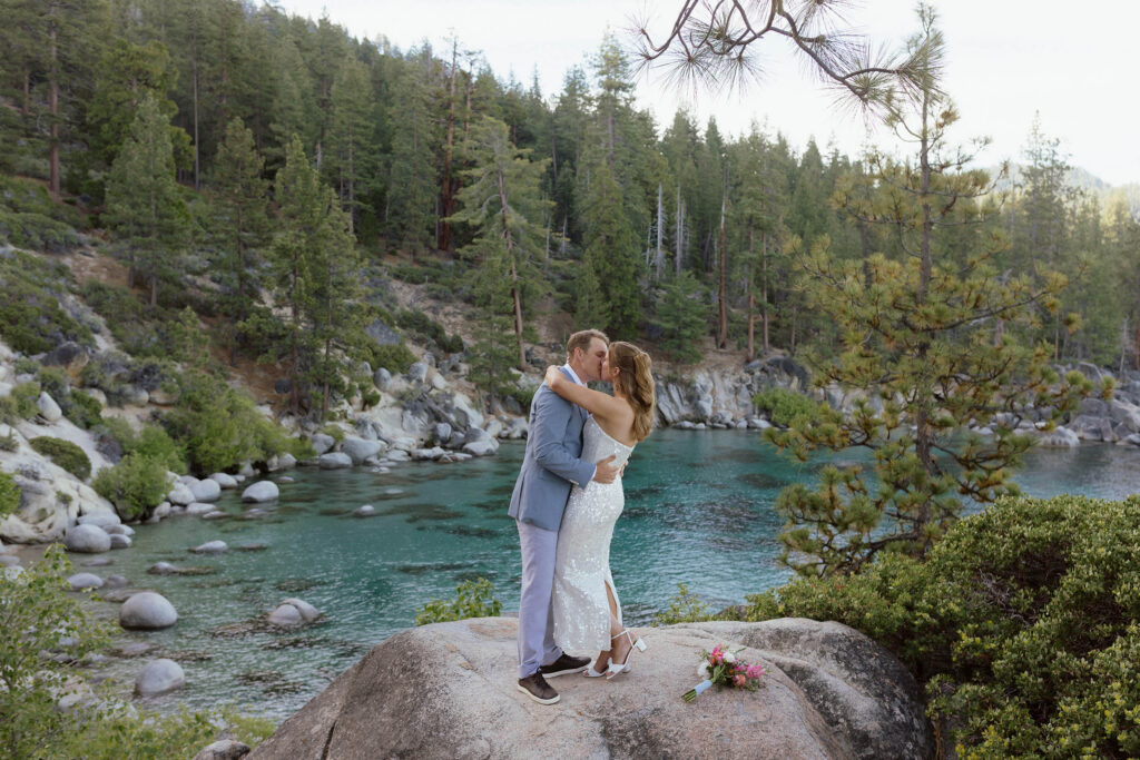 Elopement couple holding each other and kissing while standing on large boulder after ceremony with Lake Tahoe and pine trees behind them