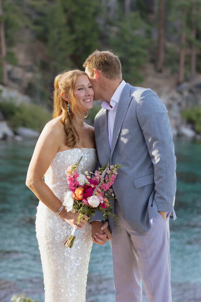 Groom whispering into bride's ear and holding her hand while she smiles and holds colorful floral bouquet with Lake Tahoe behind them