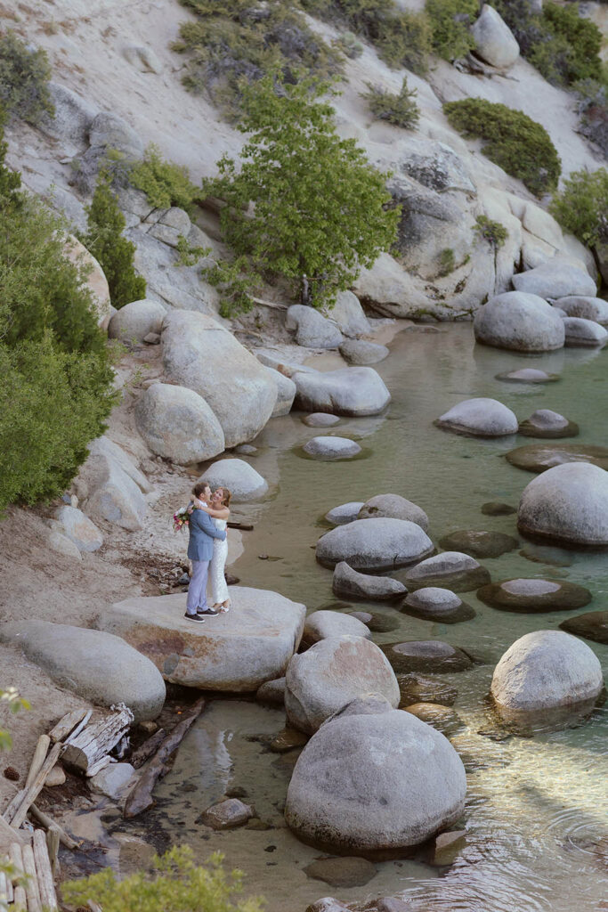 Elopement couple in the distance hugging each other while standing on top of boulder together next to Lake Tahoe