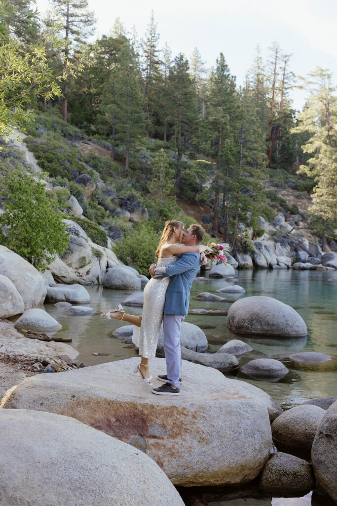 Groom hugging and lifting bride up while they kiss atop a large rock next to Lake Tahoe with pine trees in background