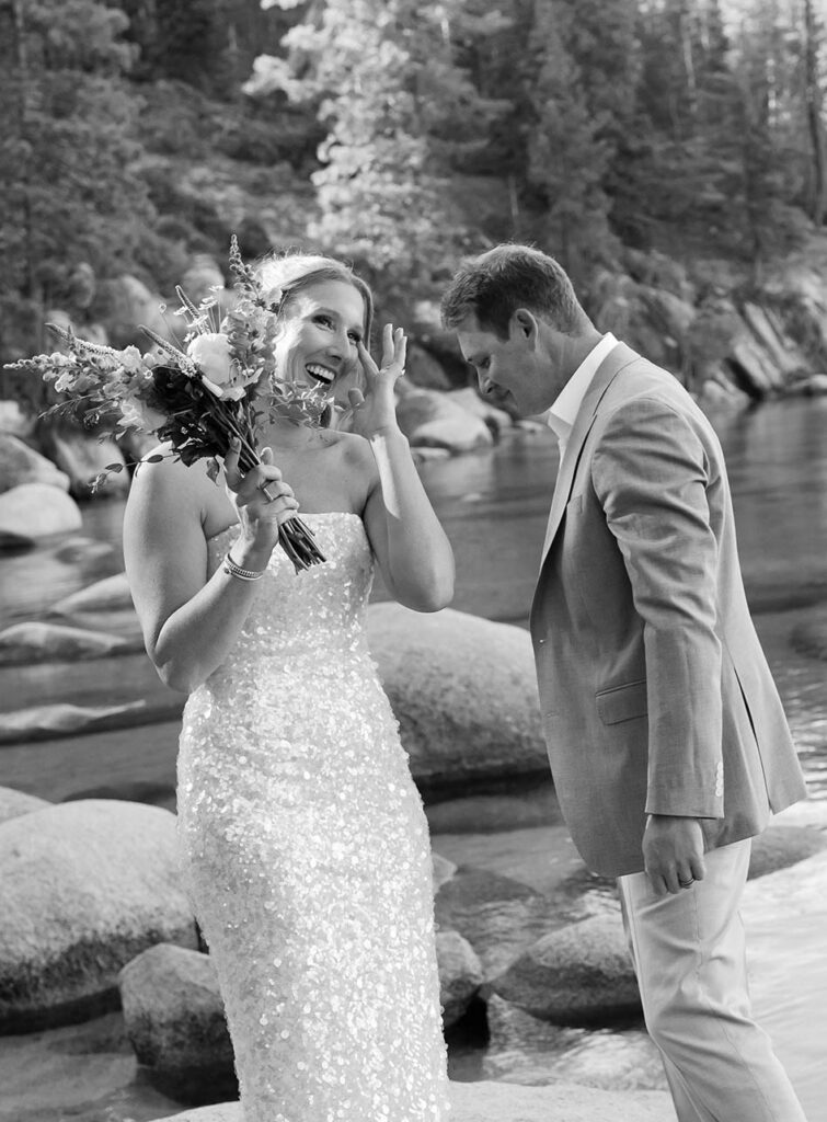 Bride laughing and wiping eye while holding floral bouquet and standing next to groom with Lake Tahoe and pine trees in background
