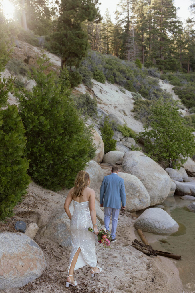 Elopement couple walking along sandy Lake Tahoe beach next to large rocks and bride holds colorful floral bouquet
