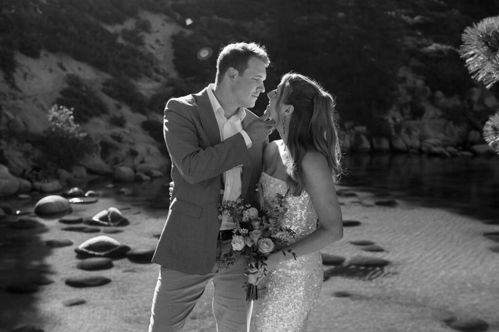 Elopement couple standing next to each other and smiling at each other while groom holds bride's chin and she holds floral bouquet with Lake Tahoe in background