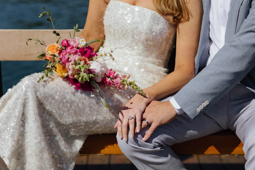 Elopement couple's hands resting on top of each other on groom's knee while wearing wedding rings and bride holds colorful floral bouquet in Lake Tahoe