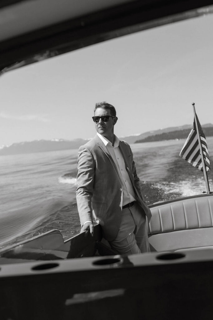 Groom leaning on side of boat with hand in his pocket and sunglasses on as the boat drives across Lake Tahoe