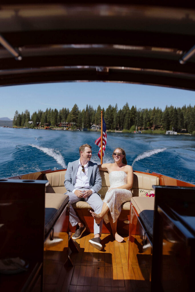 Elopement couple sitting on boat together and smiling with flag in between them while it drives across Lake Tahoe and pine trees in background
