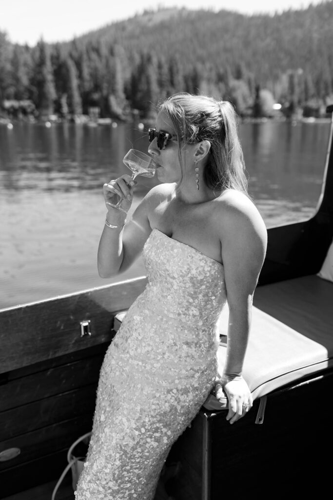 Elopement bride leaning on boat while wearing sunglasses and taking sip of drink on Lake Tahoe with pine trees in background