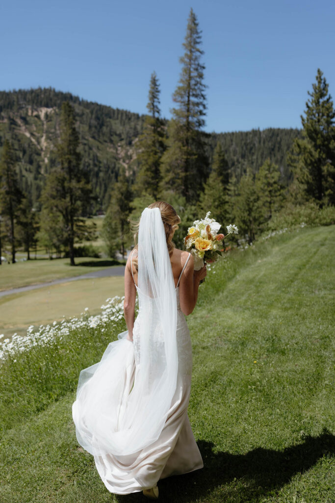 Bride in wedding dress walking across grassy field while holding colorful floral bouquet with mountains and pine trees in background at Everline Resort in Lake Tahoe