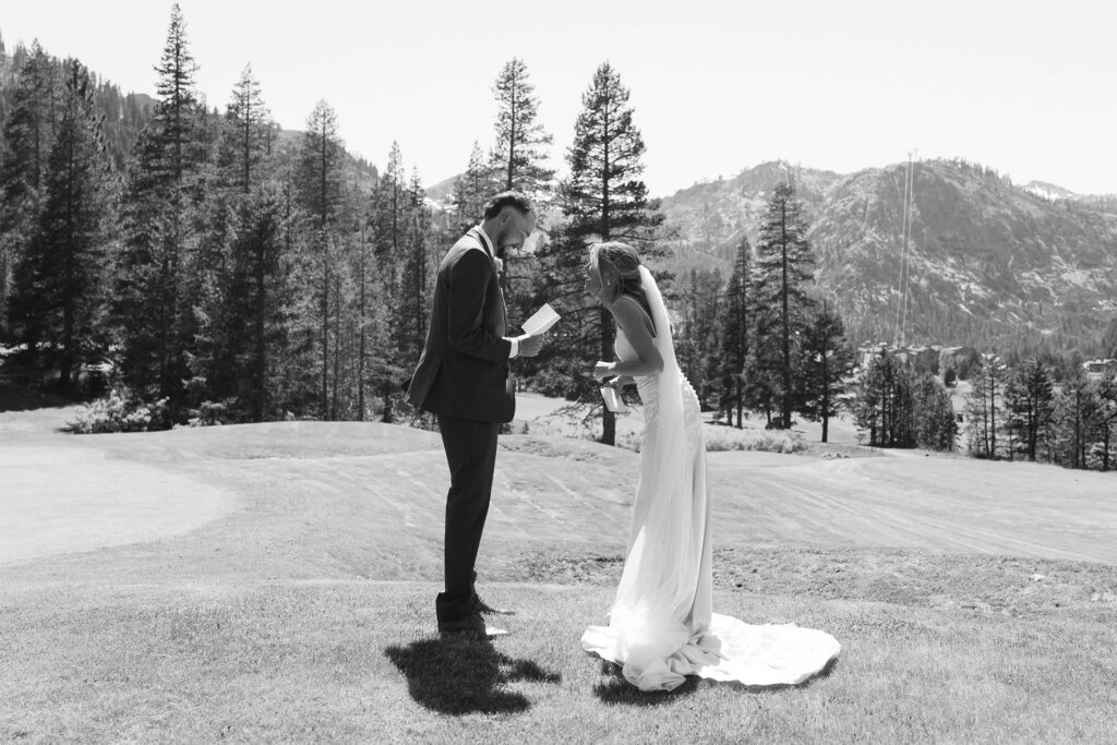 Groom reading wedding vows to bride while she laughs in an open field at Everline Resort in Lake Tahoe with pine trees and mountains in background