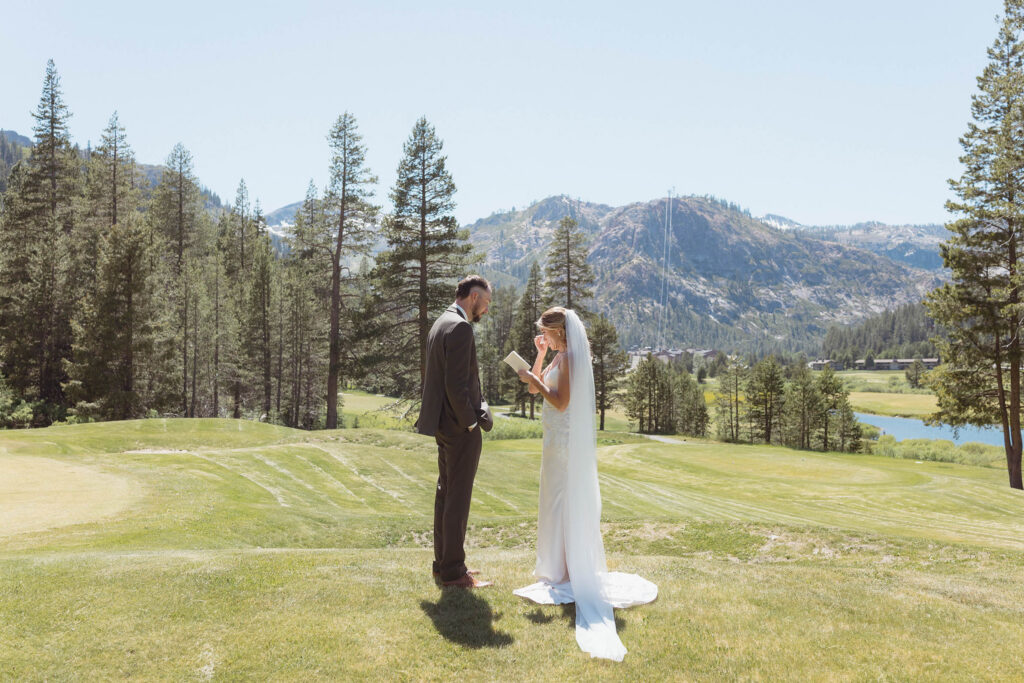 Bride reading wedding vows to groom while standing in open field with mountains and pine trees in background at Lake Tahoe