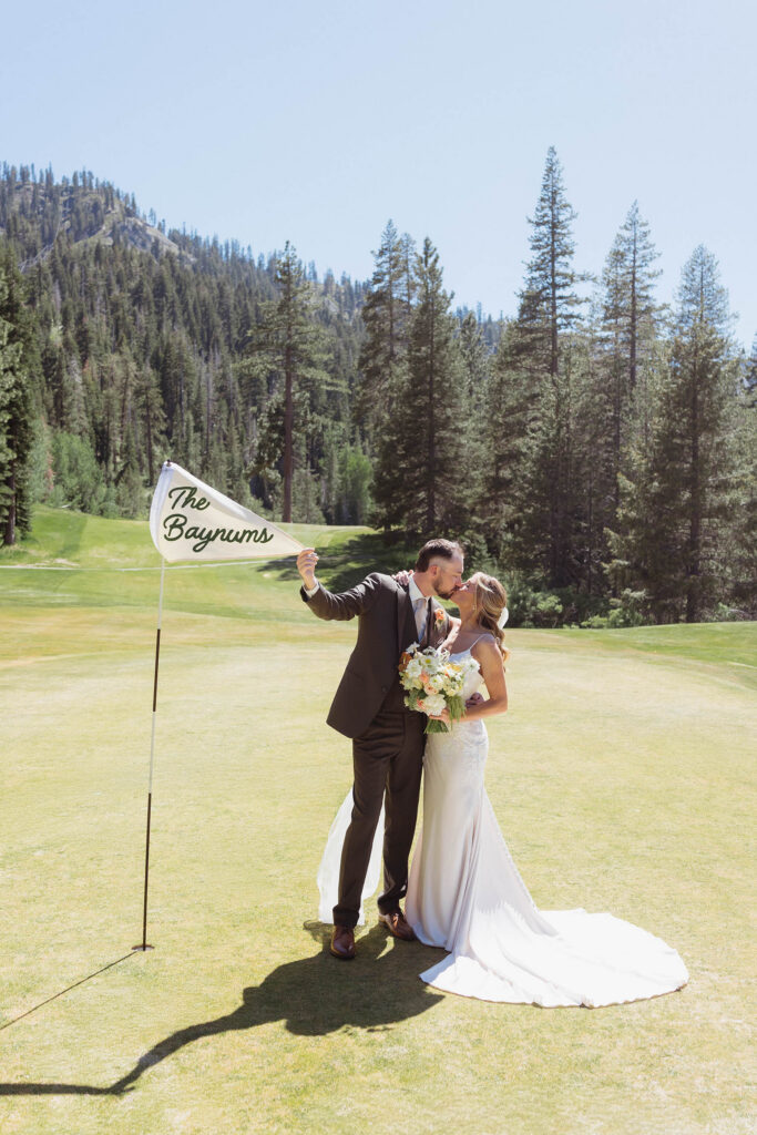 Wedding couple kissing while standing on the green of a golf course and holding flag from flag pole with their new last name at Everline Resort at Lake Tahoe