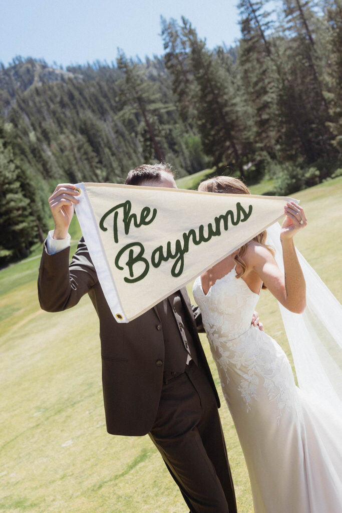 Wedding couple holding up flag with their last name while they kiss behind it at Everline Resort at Lake Tahoe with pine trees in background