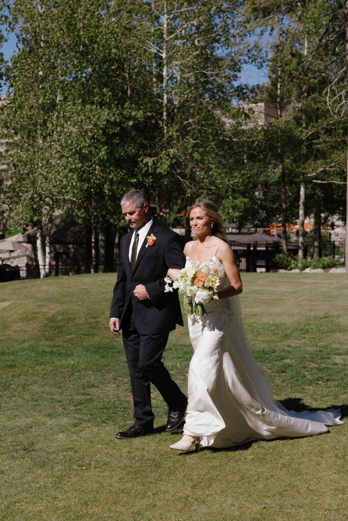 Bride holding dad's arm while walking down to wedding ceremony and holding colorful floral bouquet at Everline Resort at Lake Tahoe