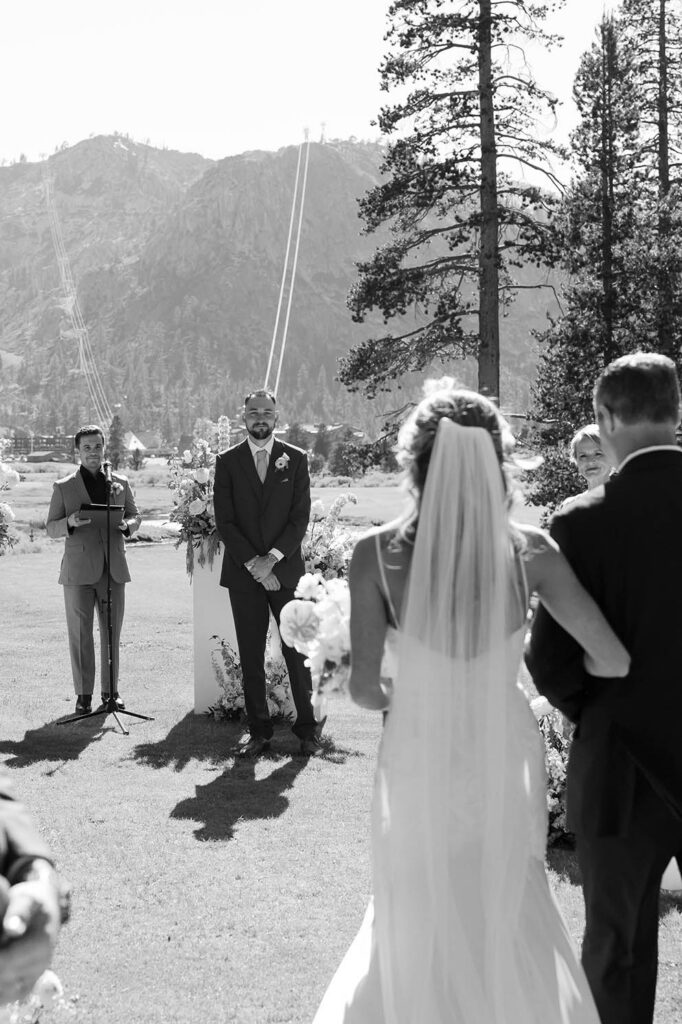 Groom smiling while watching Bride walk towards wedding ceremony holding her dad's arm at Everline Resort in Lake Tahoe with mountains in background
