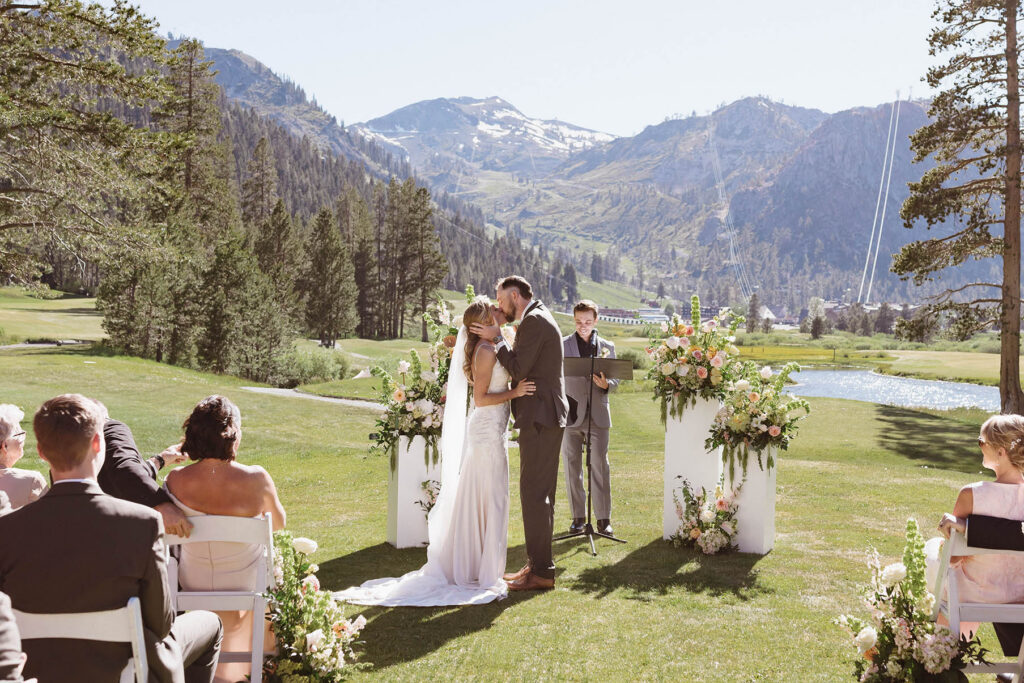 Wedding couple's first kiss during ceremony in between pillars with floral bouquets at Everline Resort at Lake Tahoe with mountains in background