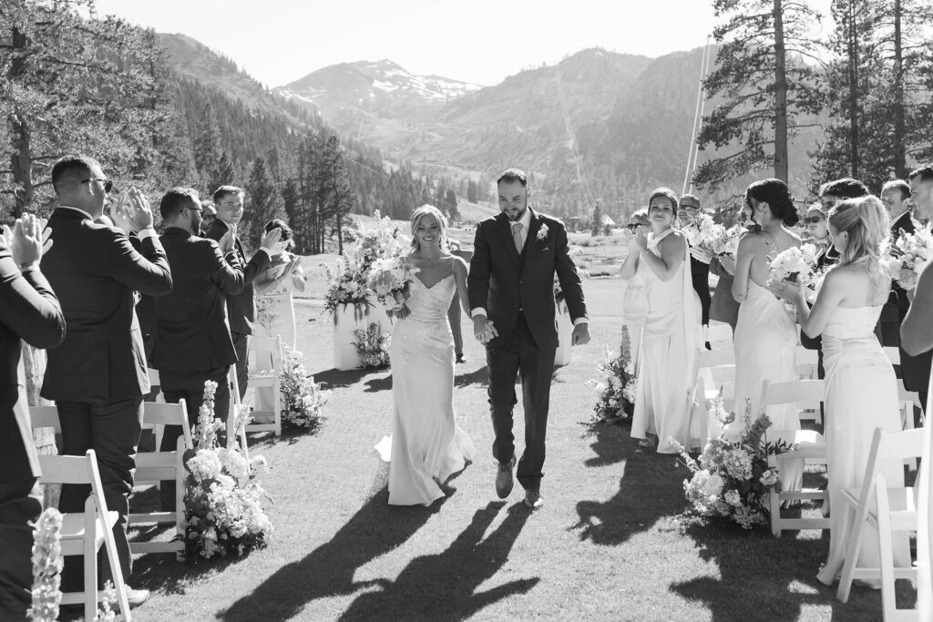 Wedding couple holding hands while walking out from ceremony at Everline Resort as guests on either side clap in Lake Tahoe with mountains in background