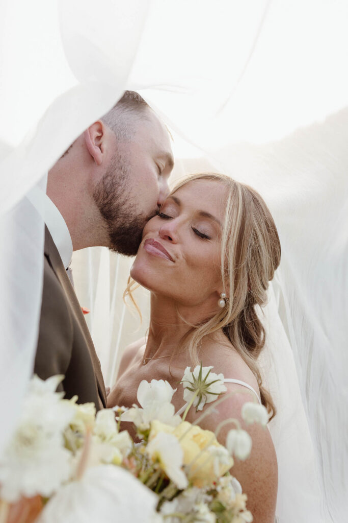 Groom kissing bride's cheek while both are underneath her wedding veil and she holds floral bouquet at Everline Resort in Lake Tahoe