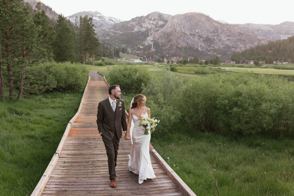 Wedding couple holding hands and walking along wooden walkway together in between lots of greenery at Everline Resort in Lake Tahoe