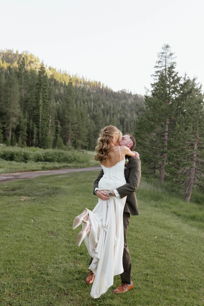 Groom picking Bride up while they kiss in an open field at Everline Resort in Lake Tahoe with forest of pine trees in background