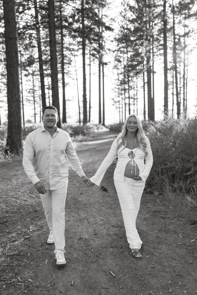 Couple holding hands while walking along trail towards camera in Lake Tahoe while woman has hand on pregnant belly