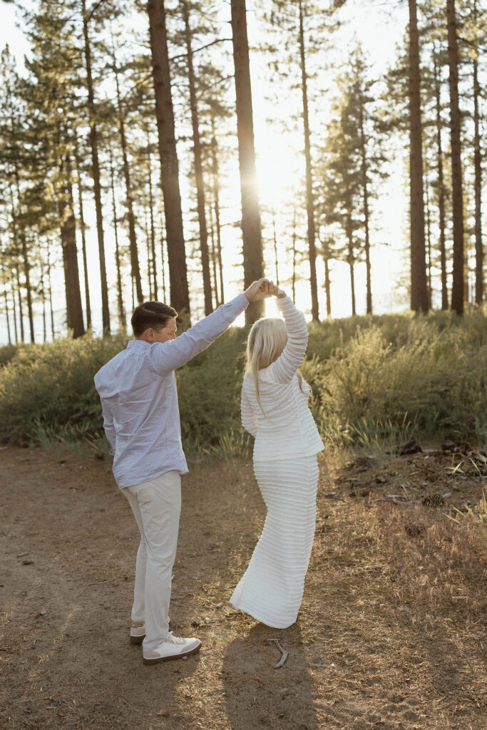 Husband twirling pregnant wife around while standing on dirt trail in Lake Tahoe with pine trees in background
