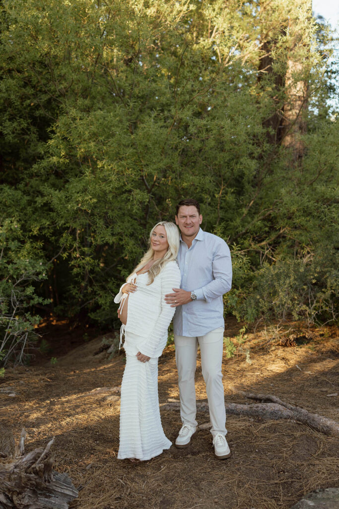 Married couple standing together and smiling at camera while in front of greenery in Lake Tahoe and wife holds her pregnant belly
