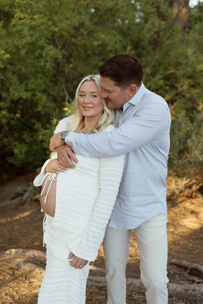 Husband hugging wife while she smiles at camera and holds her pregnant belly with greenery in background in Lake Tahoe
