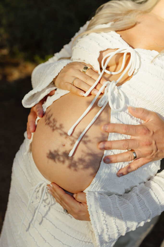 Close up of married couple's hands on pregnant wife's belly and shadows of tree branches cast over them in Lake Tahoe