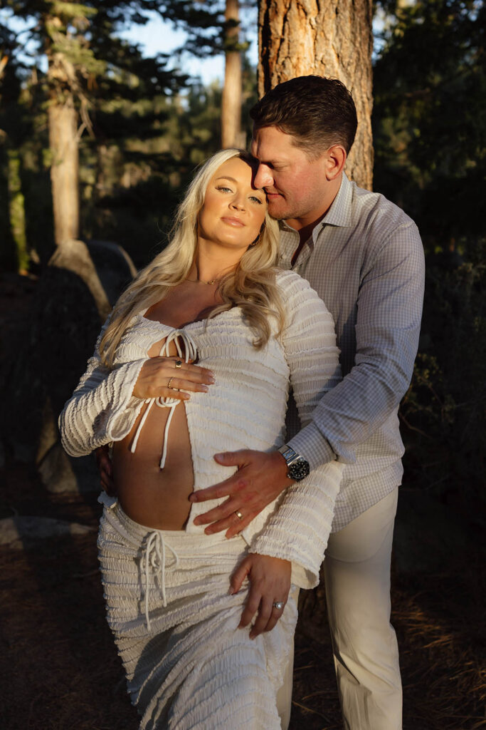 Husband hugging pregnant wife from behind while both of their hands are on her belly at sunset in Lake Tahoe