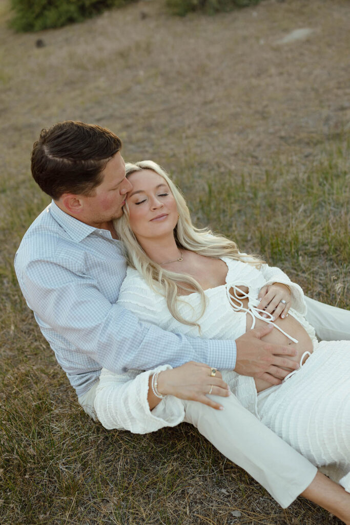 Married couple sitting together in grass while husband kisses wife's head and holds her pregnant belly in Lake Tahoe