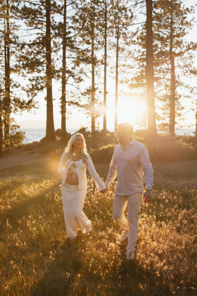 Pregnant wife and husband holding hands while walking through meadow in Lake Tahoe with sunset and pine trees in background