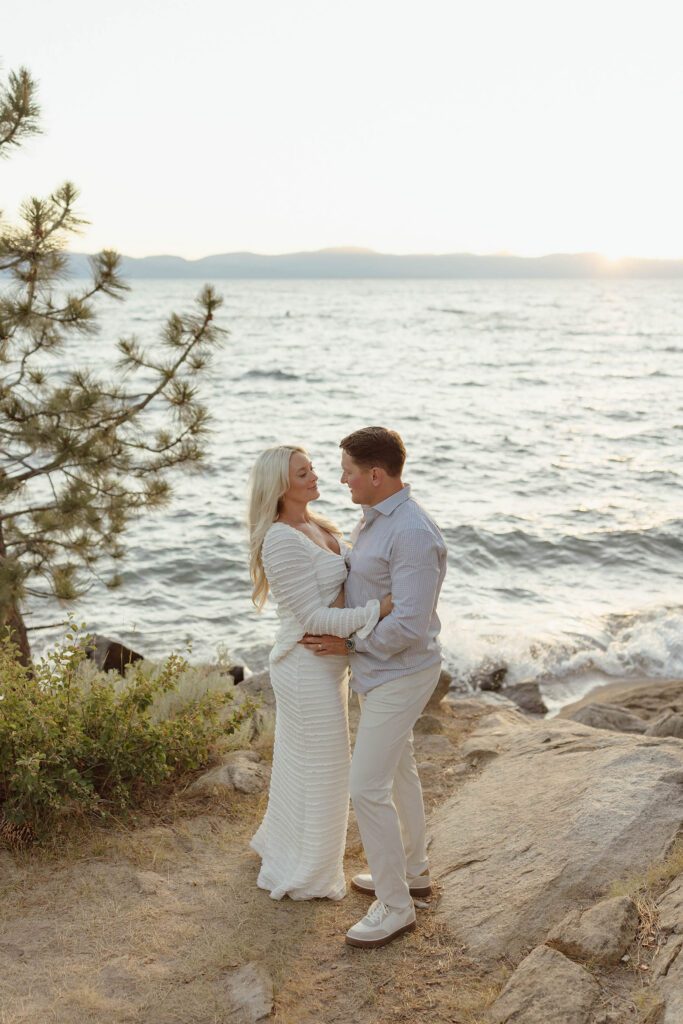 Married couple standing together on a beach in Lake Tahoe and smiling at each other while holding each other and wife is in white maternity attire