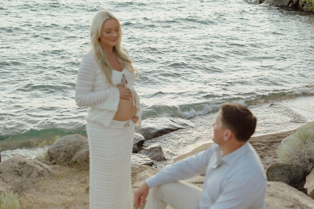 Wife holding her pregnant belly while smiling at husband sitting on a beach next to Lake Tahoe