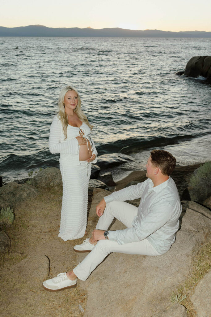 Husband sitting on rock on a beach next to Lake Tahoe and looking up at wife holding pregnant belly