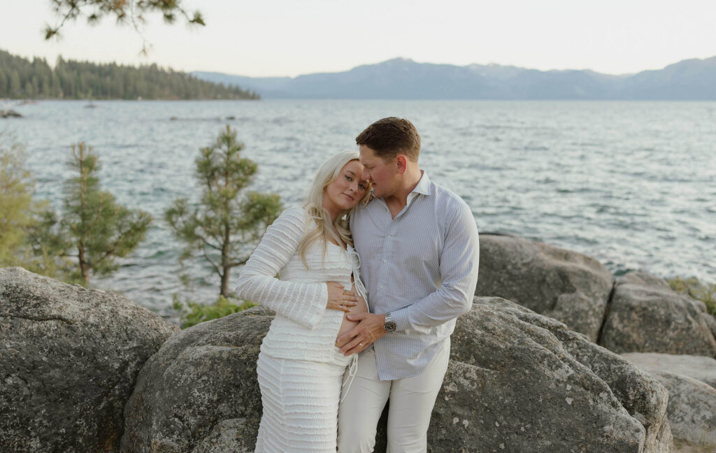 Married couple standing in front of large rock together and holding pregnant wife's belly with Lake Tahoe in background