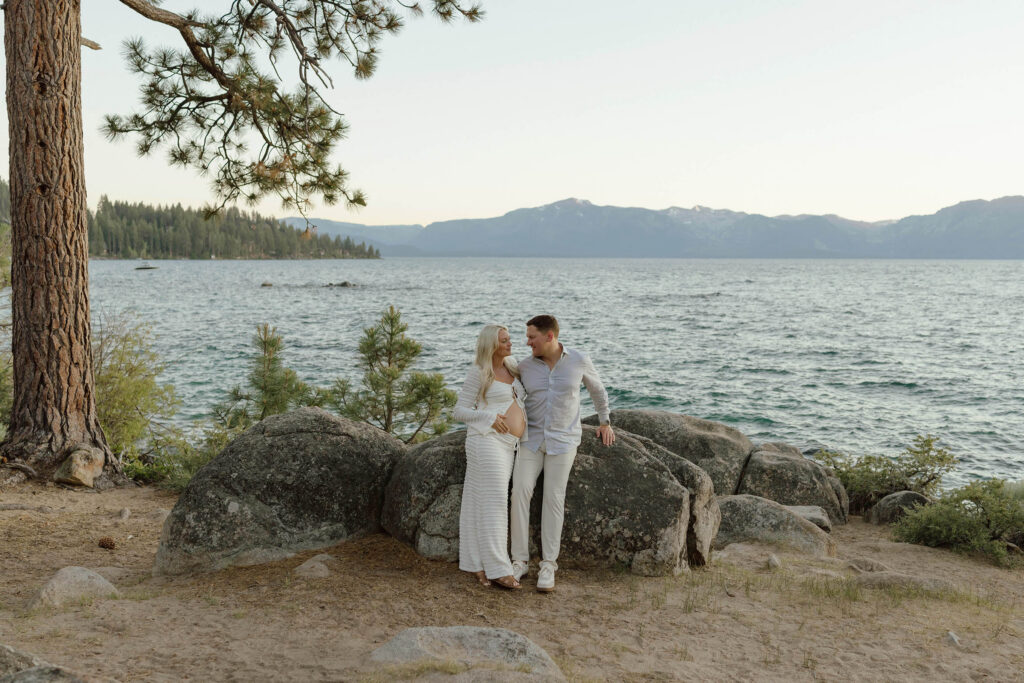 Married couple leaning on large rock together and smiling at each other while wife holds her pregnant belly with Lake Tahoe and mountains in background