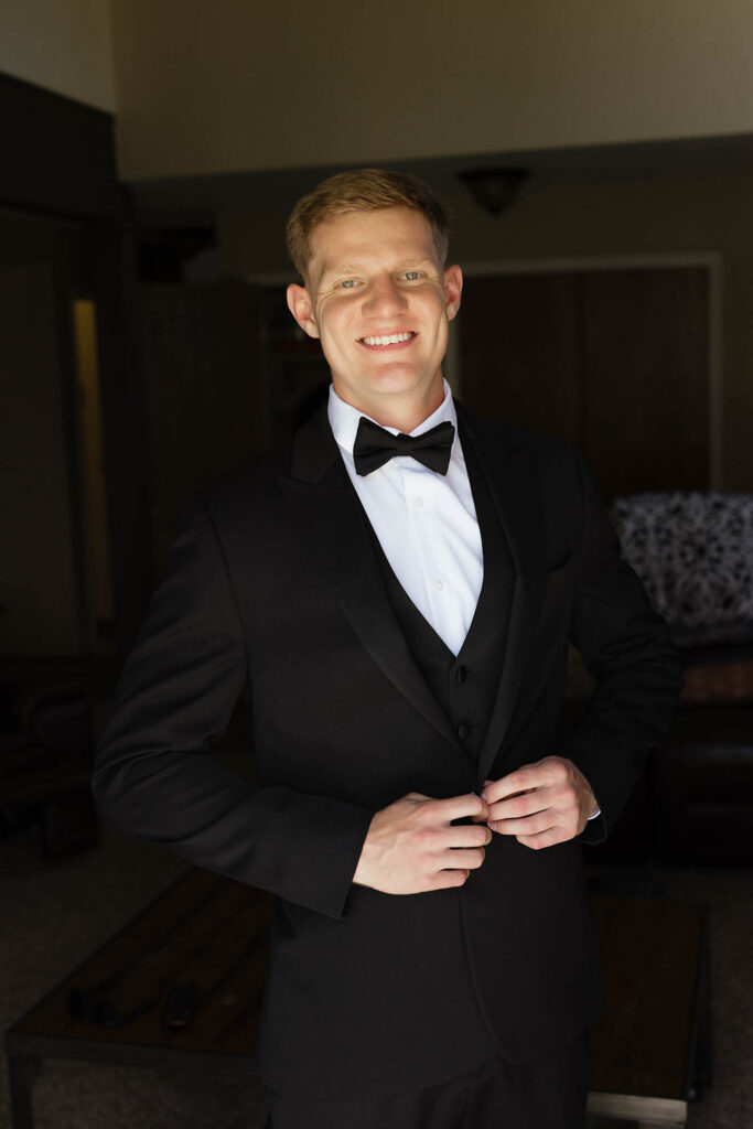 Groom smiling at camera while buttoning wedding suit jacket inside at Kinship Ranch