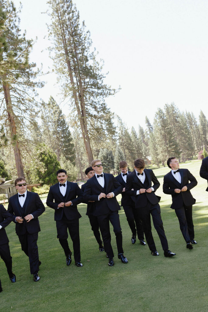 Groom and groomsmen walking across grass together while buttoning suit jackets with tall pine trees in background at Kinship Ranch