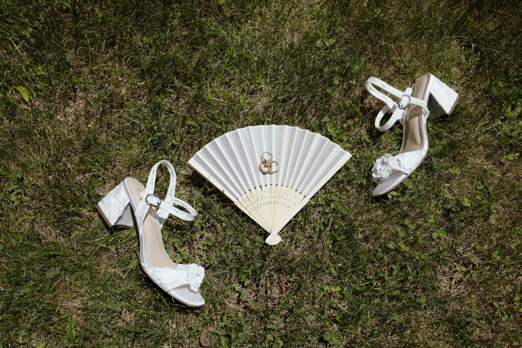 Bride's wedding rings laying on top of fan next to wedding shoes on top of grass at Kinship Ranch