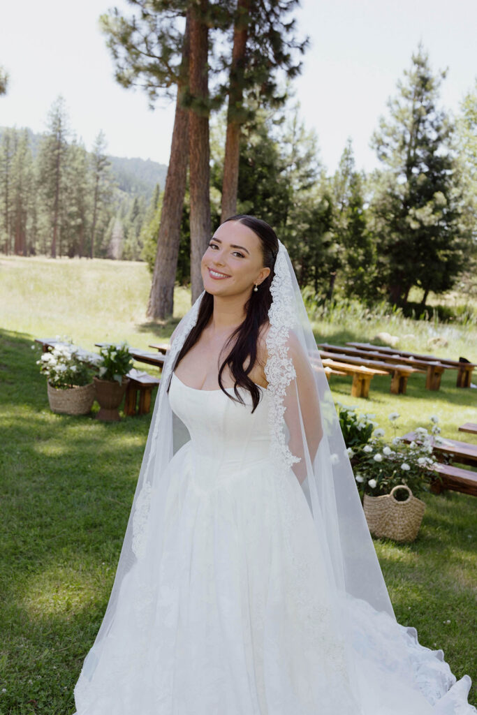 Bride smiling at camera while wearing wedding dress and veil and standing in grass in front of floral decorations and pine trees at Kinship Ranch