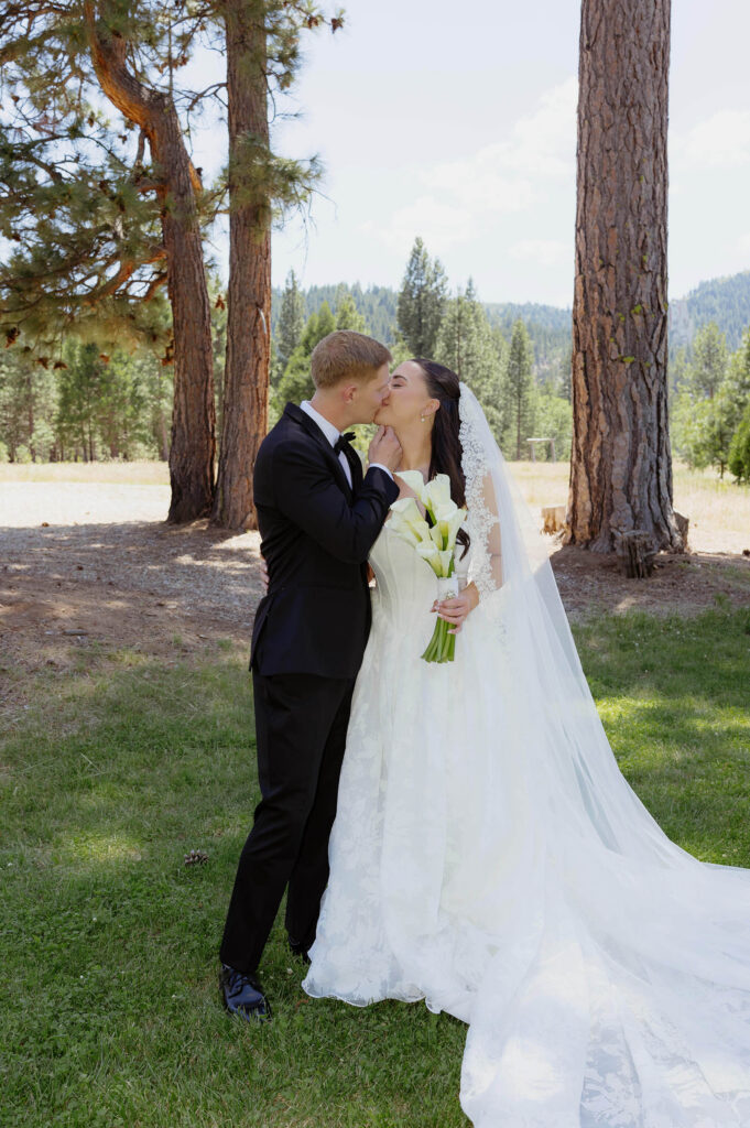 Bride and Groom kissing while in full wedding attire and bride holds white floral bouquet in front of large pine trees at Kinship Ranch