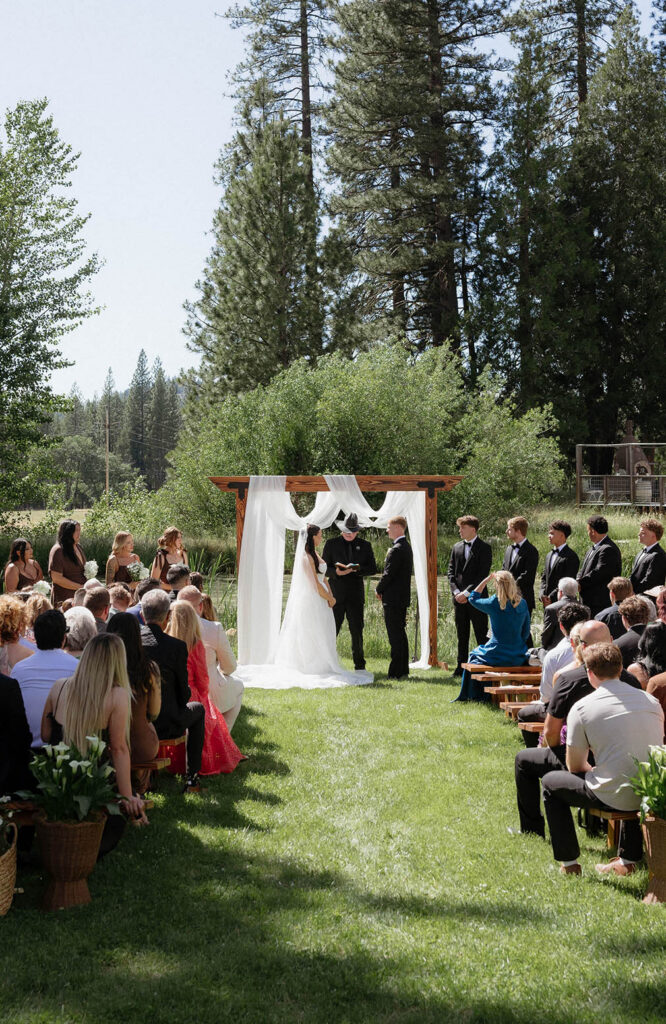 Overview of wedding ceremony at Kinship Ranch with couple standing under silk covered wooden arch and lots of greenery in background