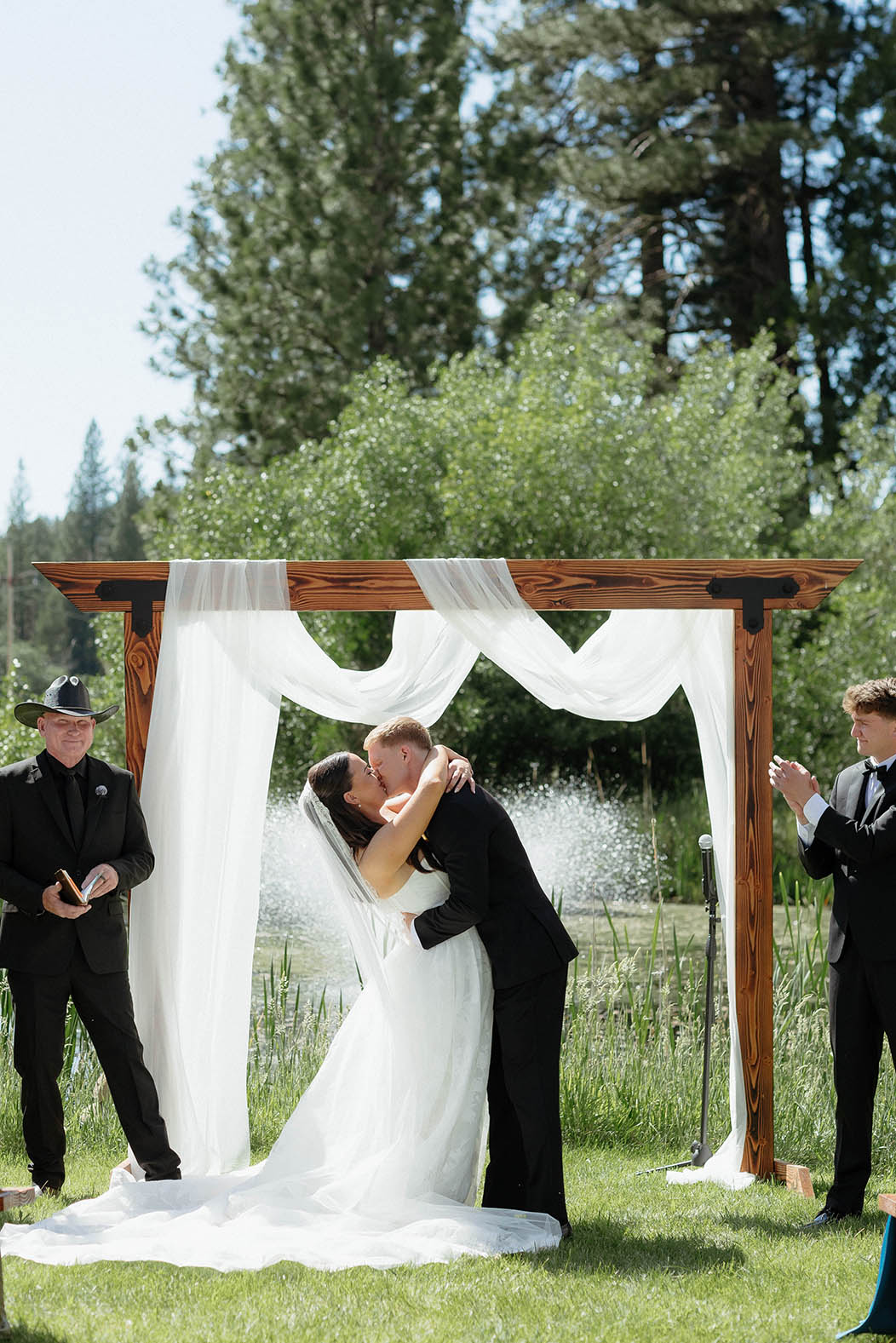 Wedding couple's first kiss after ceremony at Kinship Ranch underneath silk covered wooden arch with pine trees and greenery in background