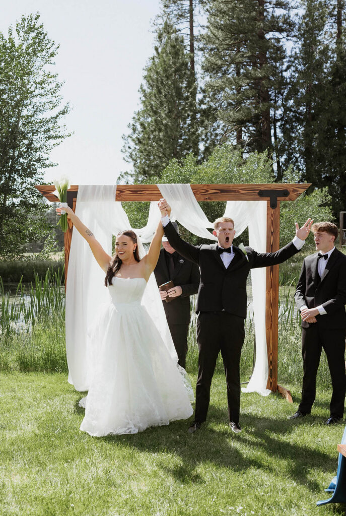 Wedding couple holding hands in the air and celebrating towards guests after ceremony at Kinship Ranch with pine trees and greenery in background